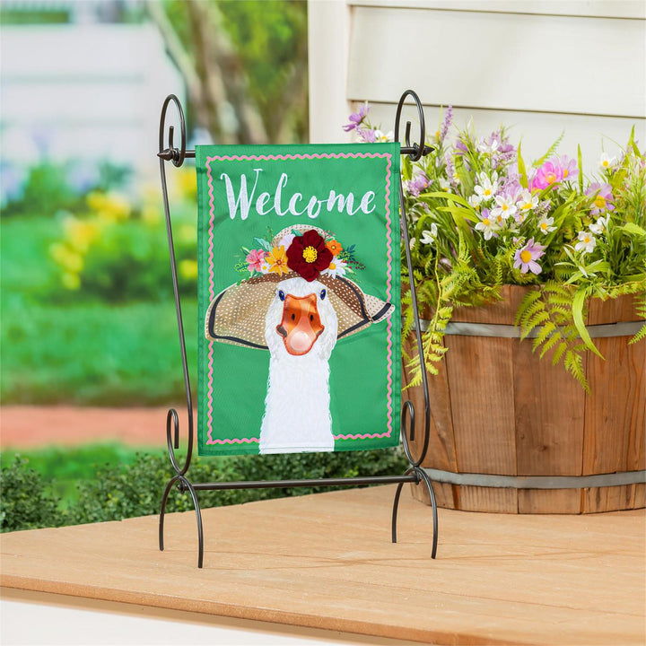Flowery Goose Welcome Garden Flag displayed on a decorative iron stand on a porch table with garden flowers