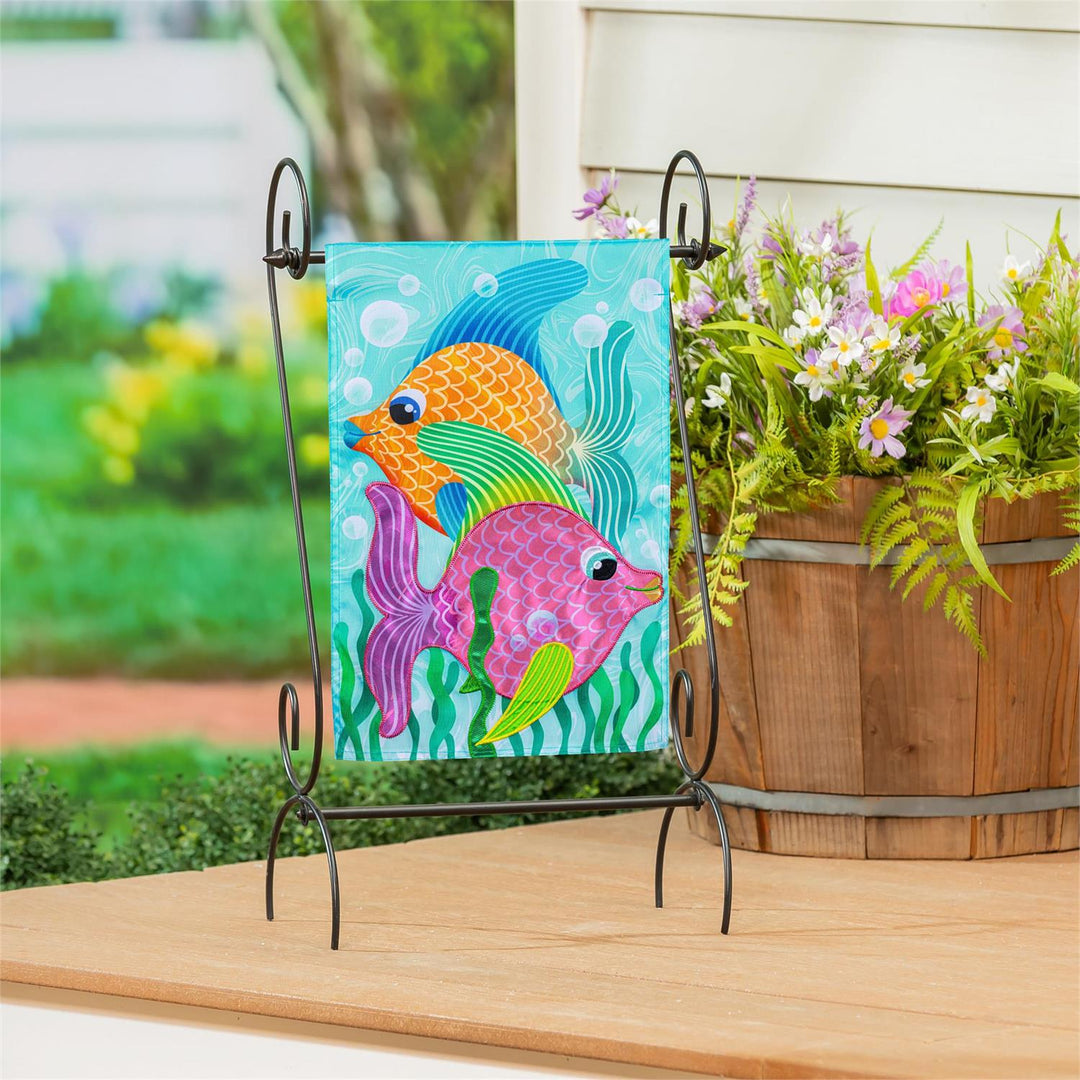 Funky Tropical Fish Garden Flag displayed on a decorative iron stand on a porch table with garden flowers