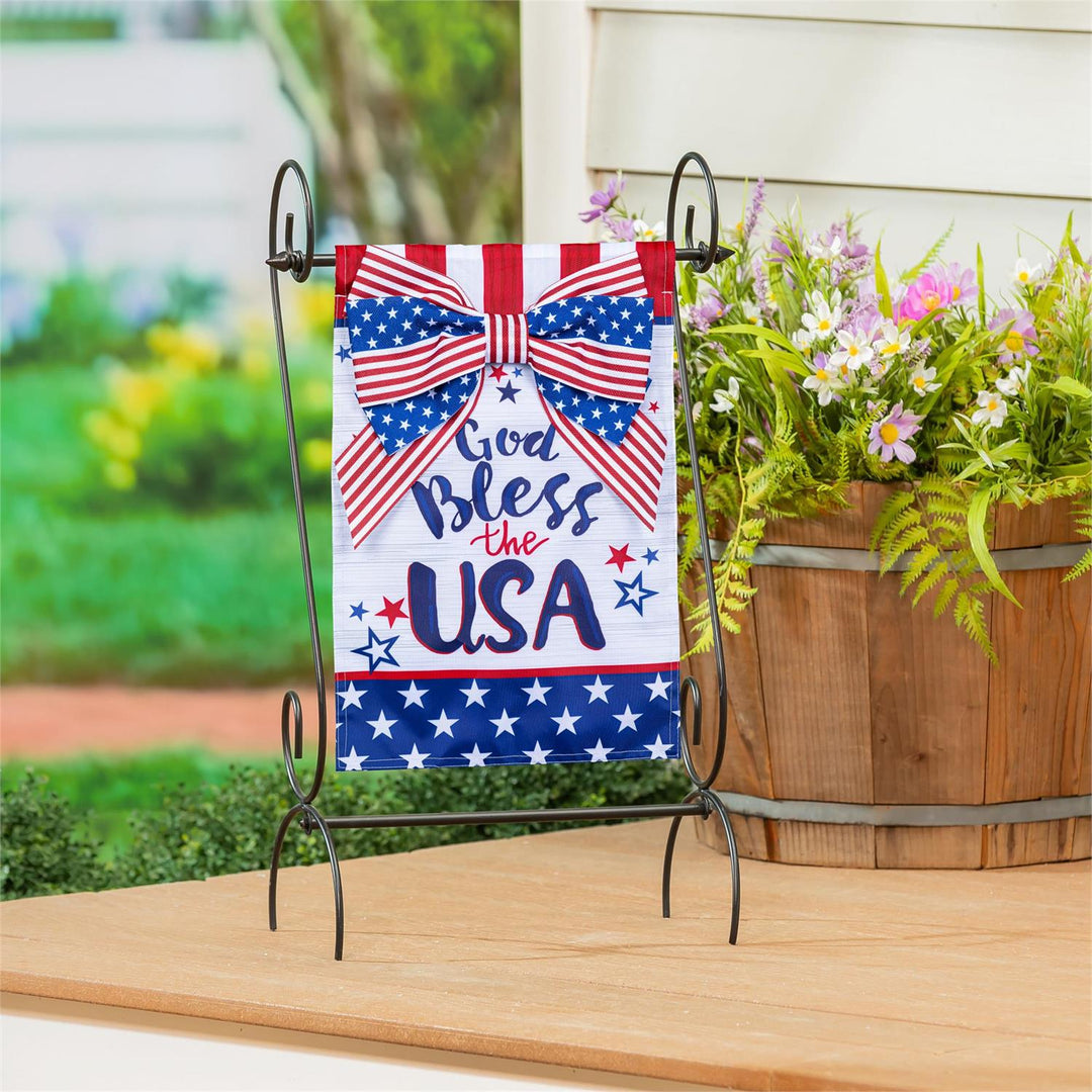 God Bless The USA Bow Garden Flag displayed on a decorative iron stand on a porch table with garden flowers