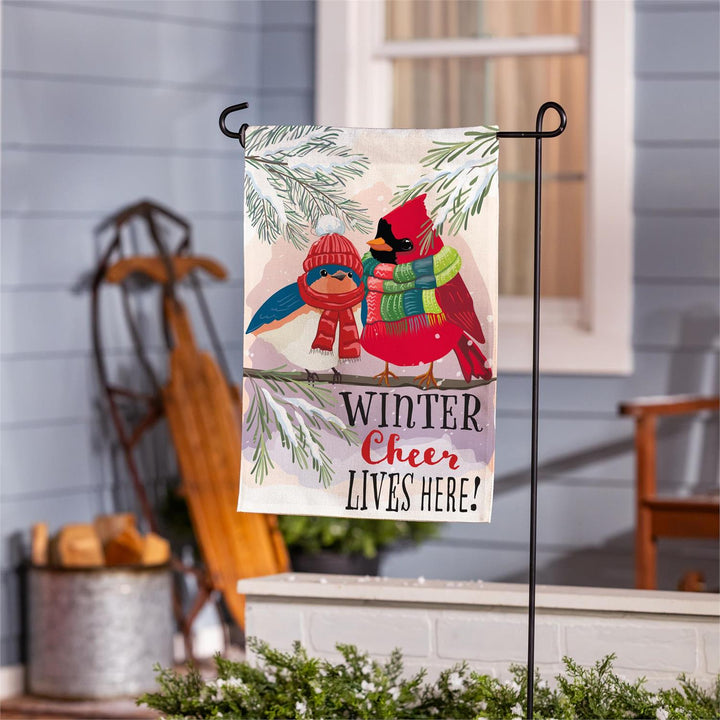 Winter Cheer bird garden flag displayed on a stand beneath snowy evergreen artwork